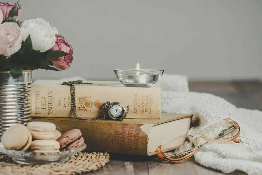 Bougie posée sur une pile de livres à coté d'une montre gousset, d'un bouquet de pivoine et quelques macarons.